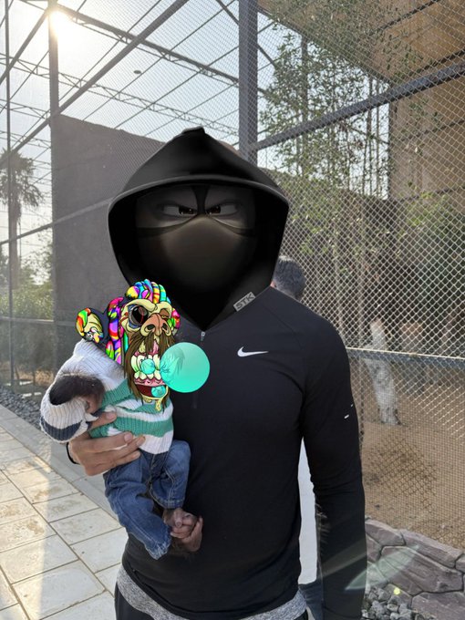 A person wearing black athletic wear with a digitally edited black geometric 'cat' or ninja-like face mask, holding a small dog wearing a colorful sombrero outfit with a teal pacifier.