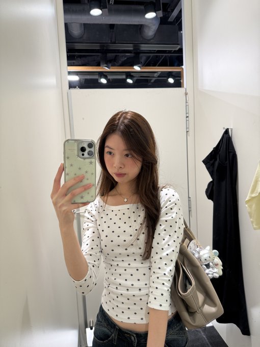 A young woman takes a mirror selfie in a fitting room, wearing a white long-sleeve top covered in black polka dots that perfectly matches the caption "polka polka." She carries a beige handbag and holds up a phone with a star-patterned case.