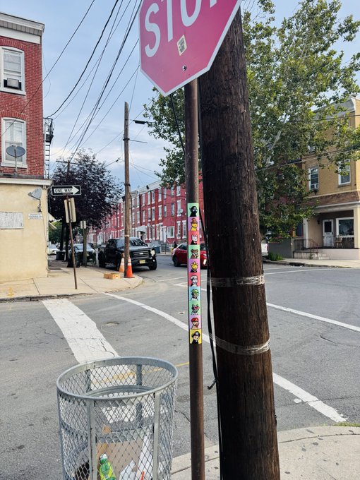 An urban street corner scene featuring a pink 'STOP' sign mounted on a wooden utility pole, with row houses, parked cars, and a wire trash can visible along the sidewalk.