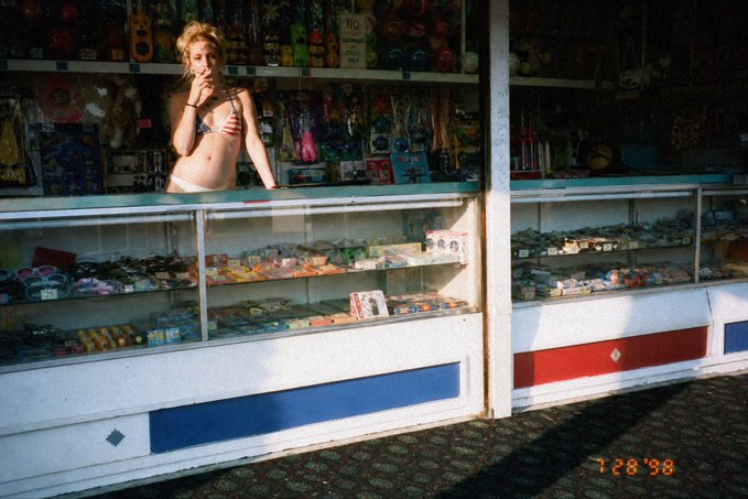 A vintage photograph from 1998 (timestamp 1/28/98) showing a young woman in a bikini standing behind the counter of a small shop or kiosk filled with merchandise.