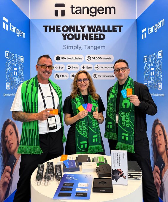Three people wearing green 'Crypto Europe Event' scarves pose at a Tangem promotional booth advertising 'THE ONLY WALLET YOU NEED' with features like '90+ blockchains' and '16,000+ assets,' displaying Tangem card products on a table.