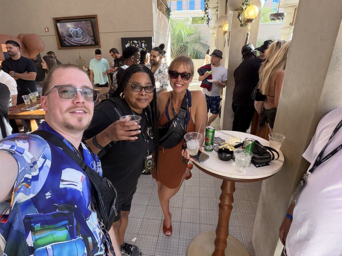 A group selfie taken at what appears to be a social event or convention, featuring people posing around a high-top table with drinks, in a venue with decorative architecture.