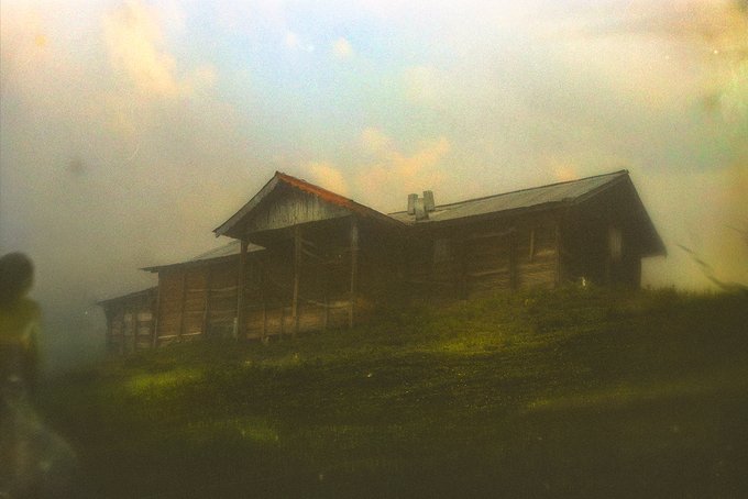 A soft-focus, vintage-toned image of a weathered wooden cabin or barn sitting on a grassy hill, with a blurred figure visible on the left edge of the frame.
