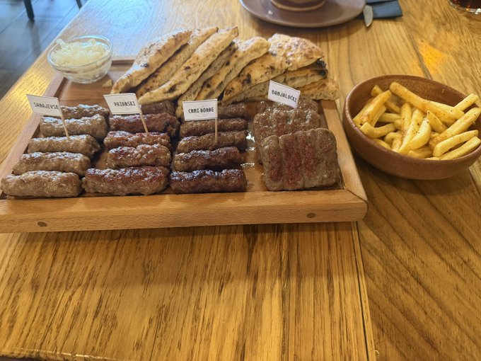 A wooden platter on a table holding an assortment of grilled sausages and flatbreads, with small labels reading 'SARAJEVSKI', 'NIŠKI', 'ČAPLJINSKI', 'DOMAĆI', and 'BJELOPOLJSKI', alongside a bowl of french fries.