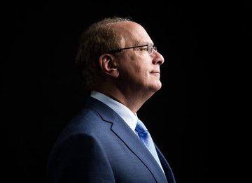 A side profile photograph shows a bald man with glasses in a blue suit, illuminated against a dark black background.