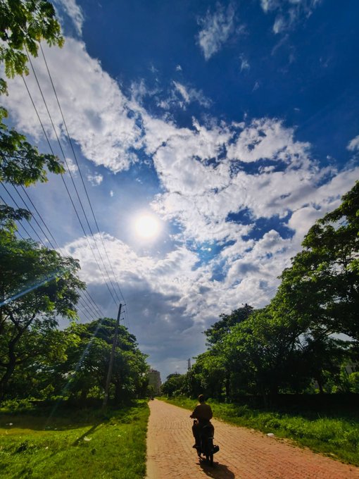 A scenic outdoor photograph showing a person riding a motorcycle down a dirt road lined with green trees under a bright blue sky with scattered clouds.