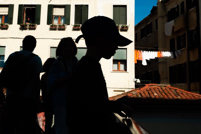 The image features a group of people walking down a street, with one person wearing a baseball cap. There are three individuals visible in the scene - two men and a woman. The man in the foreground is wearing a black hat that casts a shadow on the ground as he walks past buildings. A post on social media has been shared by someone named "Gm Ð people Happy Monday !" with an image of a person's face, likely the one wearing the baseball cap.

The background includes several buildings and clothes hanging out to dry in their windows. The photo captures the essence of everyday life in a city setting, showcasing both the urban environment and the casual attire worn by its inhabitants.
