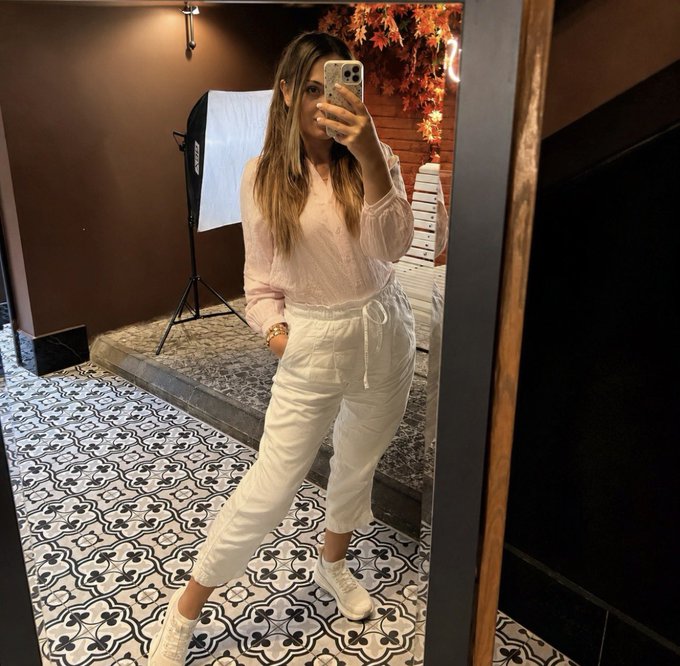 A woman taking a mirror selfie in a studio setting, wearing a sheer pink blouse, white pants, and white sneakers, with patterned tile flooring and a photography light visible behind her.