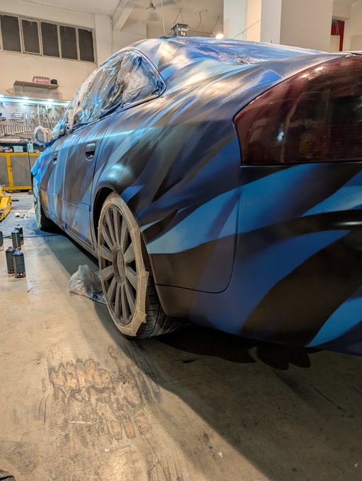 A car in a garage covered with blue and black patterned vinyl wrap, with its windows masked off during the wrapping process.