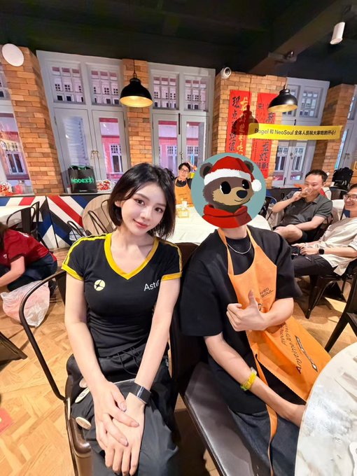 Two people sitting in a restaurant; a woman in a black and yellow 'Aster' shirt and a person wearing an orange apron with their face obscured by a teddy bear emoji filter.