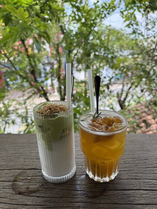 Two refreshing drinks—a layered matcha latte topped with granola and an iced amber tea—sit side by side on a wooden table against a lush green backdrop, perfectly capturing the relaxed vibe of a "good morning" weekend ritual.