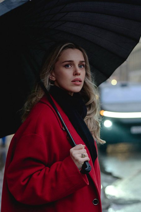 A young blonde woman in a red coat holding a black umbrella on a rainy city street.