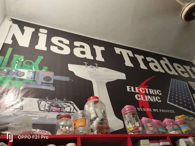 Photo taken with an OPPO F21 Pro of a storefront sign reading 'Nisar Traders' and 'Electric Clinic' above shelves displaying jars of hardware and electrical supplies.
