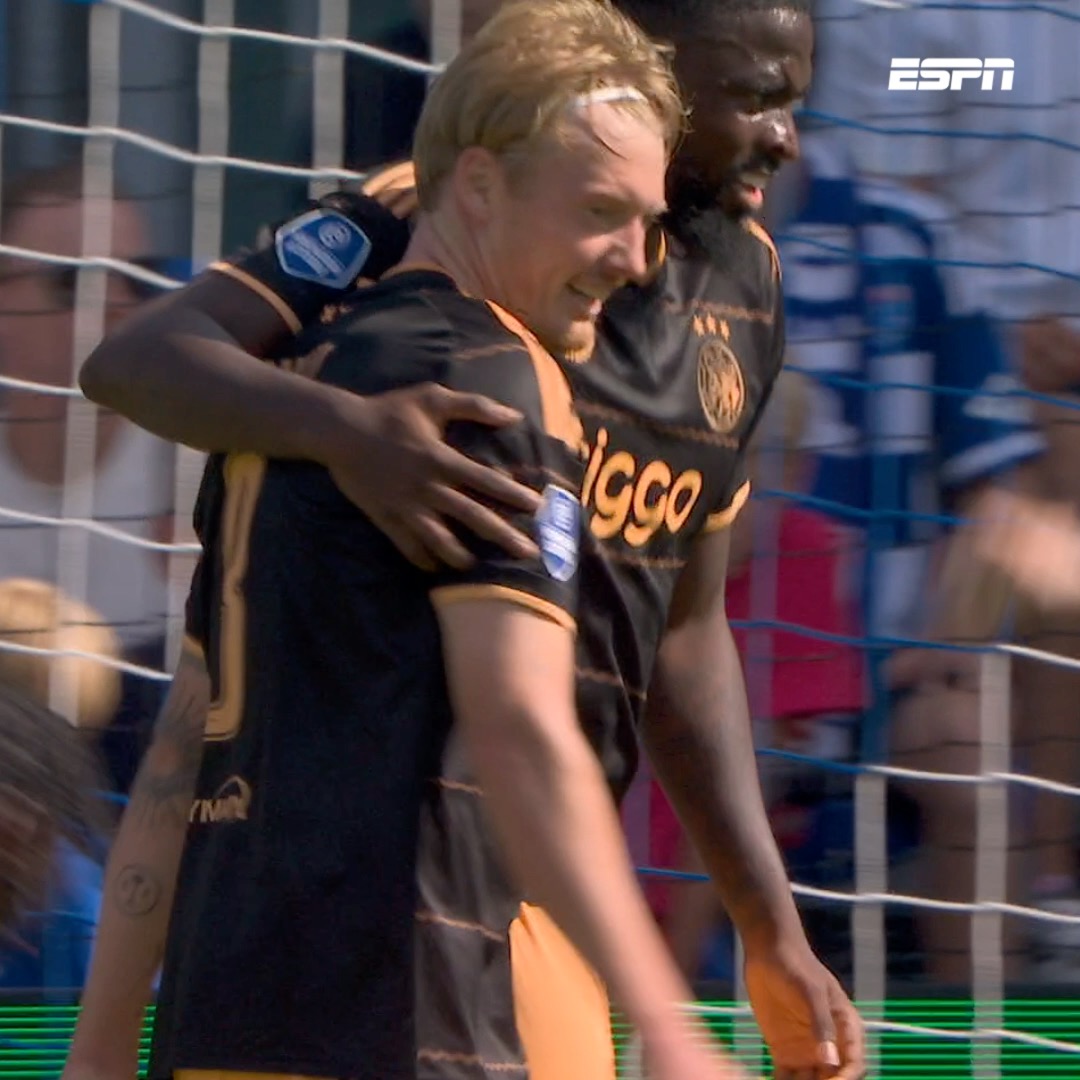 Two Ajax players, including Julian Brandt, embrace in celebration near the goal net, with their black and orange "Ziggo"-sponsored jerseys visible, capturing the joy of his debut Eredivisie goal as broadcast on ESPN.