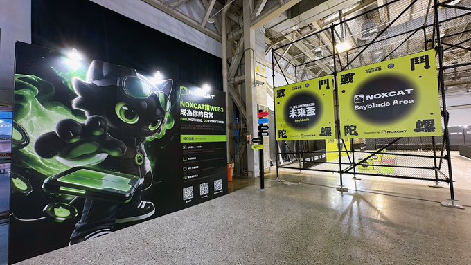An indoor event space featuring a 'NOXCAT' promotional display with a black cat mascot wearing goggles, and a 'Bayblade Area' sign in yellow with Chinese characters.