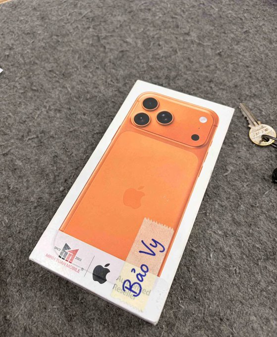 An orange iPhone box with a label reading 'Bảo Vỹ' and 'Authorized Reseller' sticker, sitting on a gray carpet next to a key.