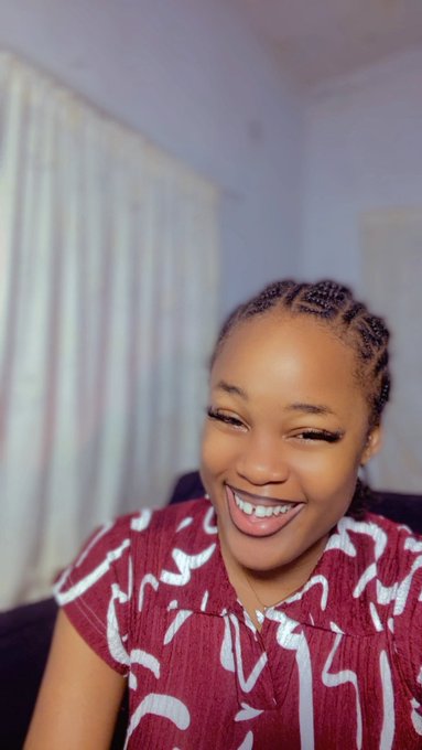A webcam-style photo of a smiling woman with braided hair wearing a maroon and white patterned top, winking at the camera in an indoor setting.