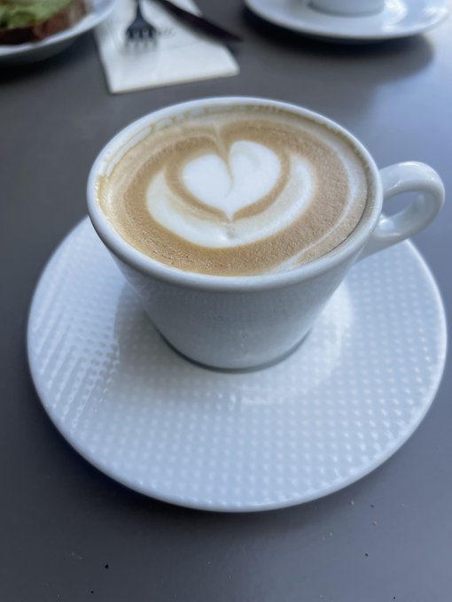 A white cup of cappuccino with a heart-shaped latte art design sits on a textured white saucer on a table.