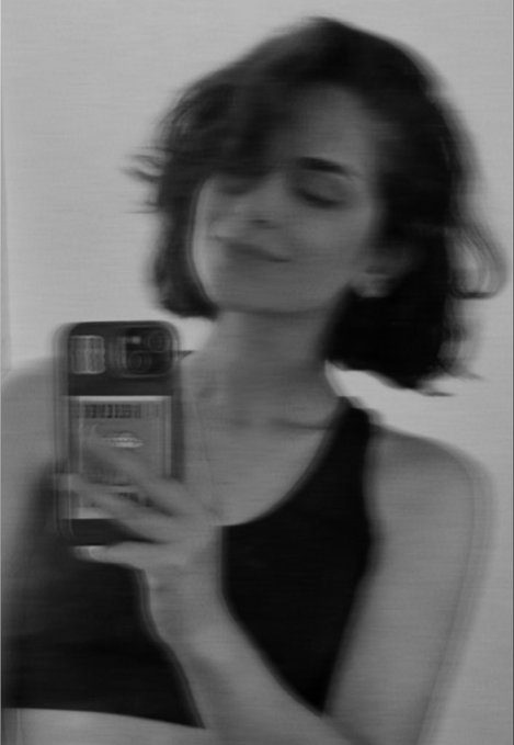 A black-and-white blurry photo of a person with short dark hair taking a mirror selfie with a smartphone, wearing a dark tank top and smiling with eyes closed.