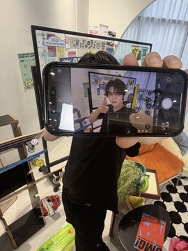 The image features a person holding up their phone, showing off an image of themselves with a green plant behind them. The photo appears to be taken at home or in a living space, as there are several books scattered around the room. The main focus is on the person and the plant, creating a sense of connection between the viewer and the subject matter.