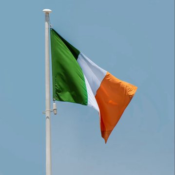 An Irish flag with green, white, and orange vertical stripes waves against a clear blue sky from a white flagpole.