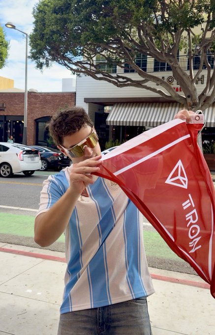 A young man wearing sunglasses and a blue and white striped shirt holds a red triangular flag with 'TRONY' text on a city street.