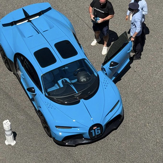 An aerial view of a light blue Bugatti hypercar with the number '16' on it, with two men standing beside the open door on a paved surface.