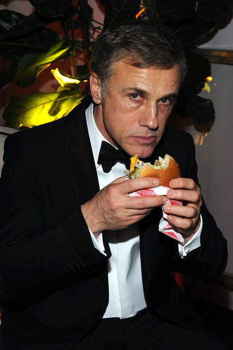 A man in a black tuxedo with a bow tie eats a hamburger while holding it with a napkin.