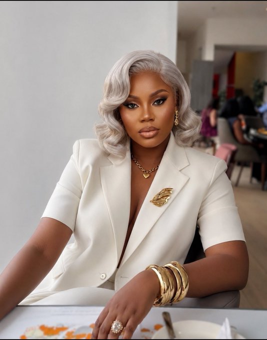 A portrait of a woman with silver hair wearing a white blazer adorned with gold jewelry, including a leaf brooch, chain necklace, and stacked bangles, seated in a restaurant setting.