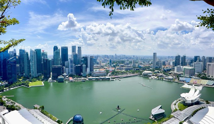 A sweeping aerial view of Singapore's Marina Bay, featuring the iconic skyline, towering financial district skyscrapers, and the lotus-shaped ArtScience Museum along the waterfront under a bright, cloud-filled sky. The post humorously references the city's status as a major crypto conference hub, where attendees endure long-haul flights to network, fueled by nicotine pouches and ambition.