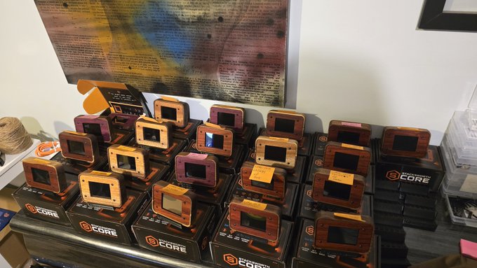 A workbench display showcases 21 handcrafted "21 STACKSWORTH COREs" — wooden-framed Bitcoin hardware devices with small screens, arranged in rows atop branded boxes labeled "21 CORE BY 21.STACKSWORTH." The image visually corresponds with the post's announcement of a completed production run, with each unit built, flashed, and tested ahead of the Vancouver Bitcoin Block Party.