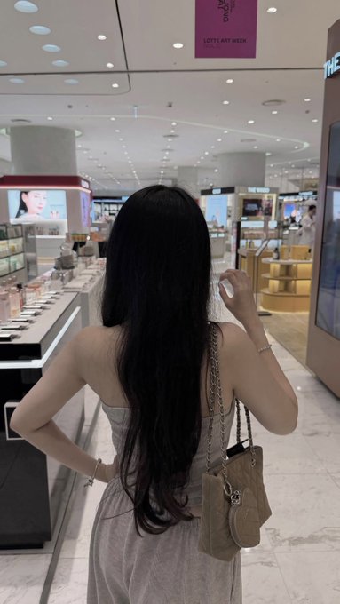 A photo of a woman from behind with long black hair, wearing a grey top and carrying a brown handbag, standing in a cosmetics store with a 'LOTTE ART WEEK' sign visible above.