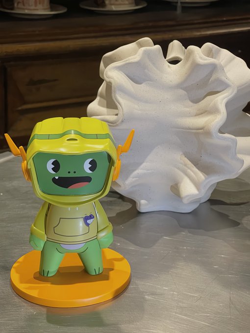 A cute green frog-like astronaut figurine in a yellow spacesuit stands on a bright orange base next to a large white coral sculpture, giving off cheerful "Gm world" vibes to start the day.