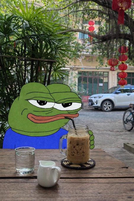 A cartoon Pepe the Frog sits at a wooden outdoor café table sipping iced coffee through a straw, with a glass of water and small white pitcher nearby, set against a backdrop of lush greenery and red lanterns. The relaxed, peaceful scene matches the post's calm "gm" (good morning) vibe, celebrating the simple joy of a quiet, blessed morning coffee moment.