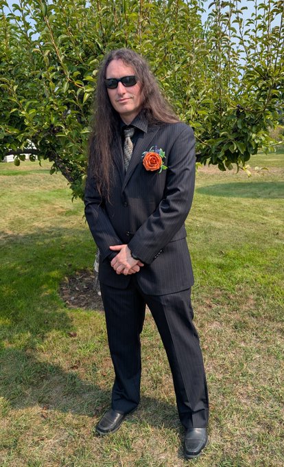 A man with long dark hair and sunglasses wearing a black pinstripe suit with a red rose boutonniere, standing outdoors in front of a green bush.