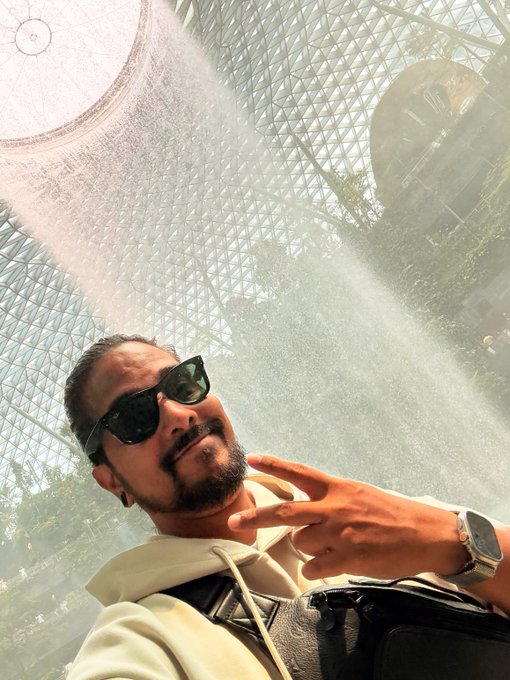 A man with a beard and sunglasses takes a selfie under the indoor waterfall at Jewel Changi Airport in Singapore, matching the "GM Singapore" post by showcasing one of the city's iconic landmarks.