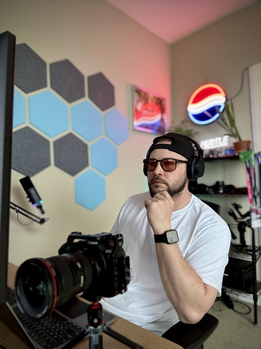 A man wearing glasses, a backwards cap, and headphones sitting at a desk with a professional camera in front of him, in a room with hexagonal acoustic panels and a Pepsi neon sign.