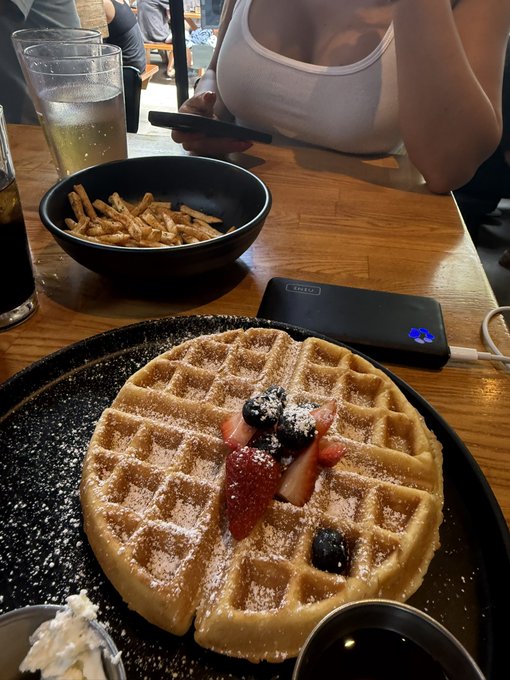 A restaurant meal featuring a large waffle topped with strawberries, blueberries, and powdered sugar on a black plate, served alongside a bowl of fries and drinks on a wooden table.