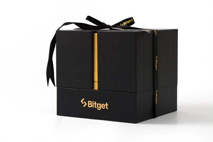 A black Bitget-branded gift box tied with a black ribbon and gold accent stripe, containing the exclusive "Golden Ball" merchandise awarded to two lucky Universal Cup winners alongside the $350,000 prize pool. The Bitget logo is prominently displayed in gold on the front of the box, matching the premium merch prize mentioned in the post.