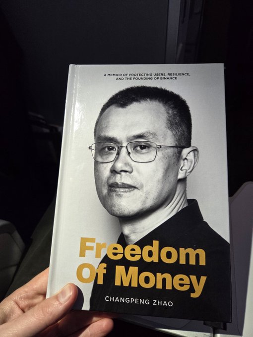 A hand holds a book titled 'Freedom Of Money' by Changpeng Zhao, subtitled 'A memoir of protecting users, resilience, and the founding of Binance,' featuring a black and white cover photo of the author.