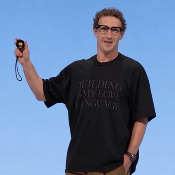 A man with glasses and curly hair stands on a stage against a blue background, holding up a small black device. He wears a black t-shirt reading 'BUILDING IS MY LOVE LANGUAGE'.