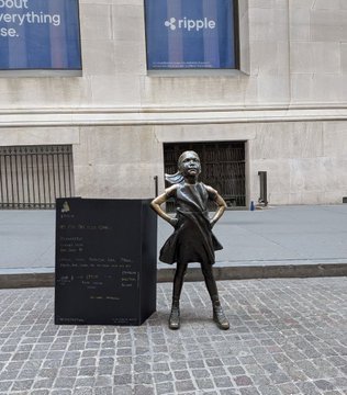 The image features a bronze statue of a girl standing next to a black sign with white text, which says "market's down to sh*t but she' s not moving! neither are we onwards and upwards epochprotocol". The girl is dressed in a dress and has her hands on her hips. The scene takes place outside a building, possibly an office or a store.