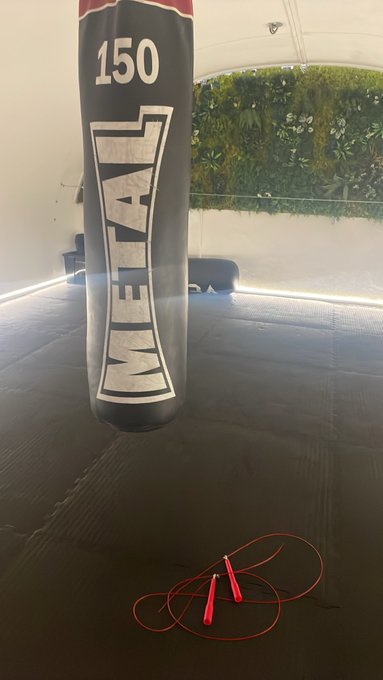 A black 'METAL' punching bag with '150' on it hanging in what appears to be a tent or covered outdoor area, with a red jump rope on the floor below.
