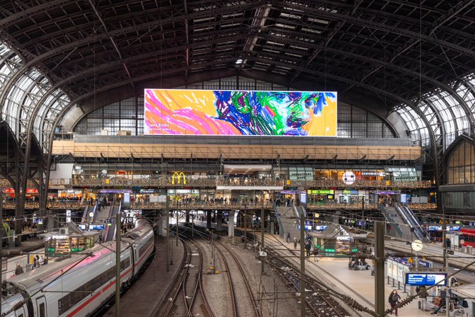 The image shows a large, colorful billboard inside an airport terminal with multiple levels of platforms and escalators. A train can be seen passing by under the billboard, adding to the bustling atmosphere of the scene. The text "GM you may want to apply for this" is present in the image as well.