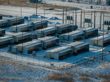 An aerial view of a snow-covered industrial facility with multiple rectangular buildings, electrical equipment, and a chain-link fence under winter conditions.