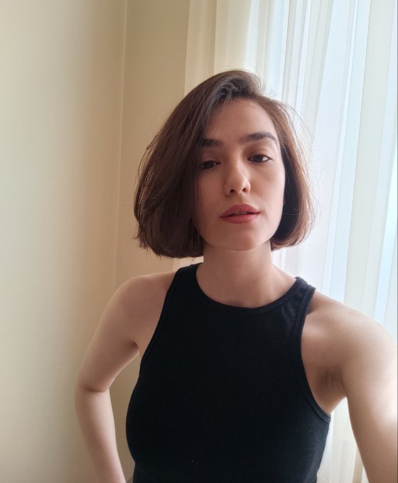 A selfie-style portrait of a young woman with a short brown bob haircut wearing a black sleeveless top, standing near sheer white curtains by a window.
