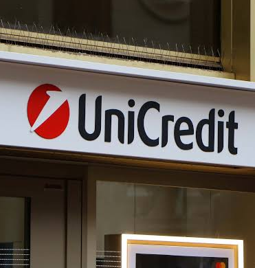 The image shows the exterior signage of UniCredit bank, featuring its distinctive red logo and bold black lettering on a white background. This visual connects directly to the news of Italy's second-largest bank exploring crypto custody and brokerage services, highlighting the financial institution at the center of the development.