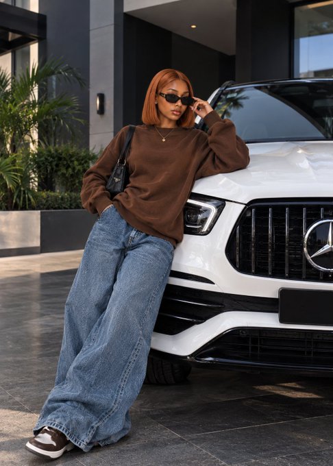 A woman with a red bob haircut wearing sunglasses, a brown sweatshirt, and baggy jeans poses leaning against a white Mercedes SUV.