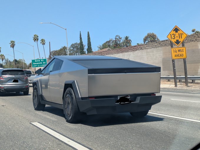 A silver Tesla Cybertruck is seen from behind driving on a highway, with a yellow road sign indicating '13'-11"' and '1/4 MILE AHEAD,' along with a green exit sign for 'Boto St.'