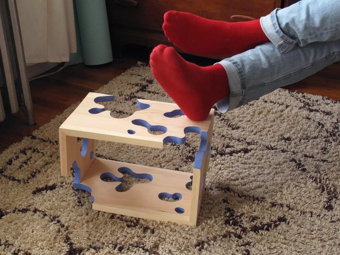 A person's feet in red fuzzy socks are propped up on a small wooden step stool with a blue swirl pattern, resting on a textured beige shag rug.